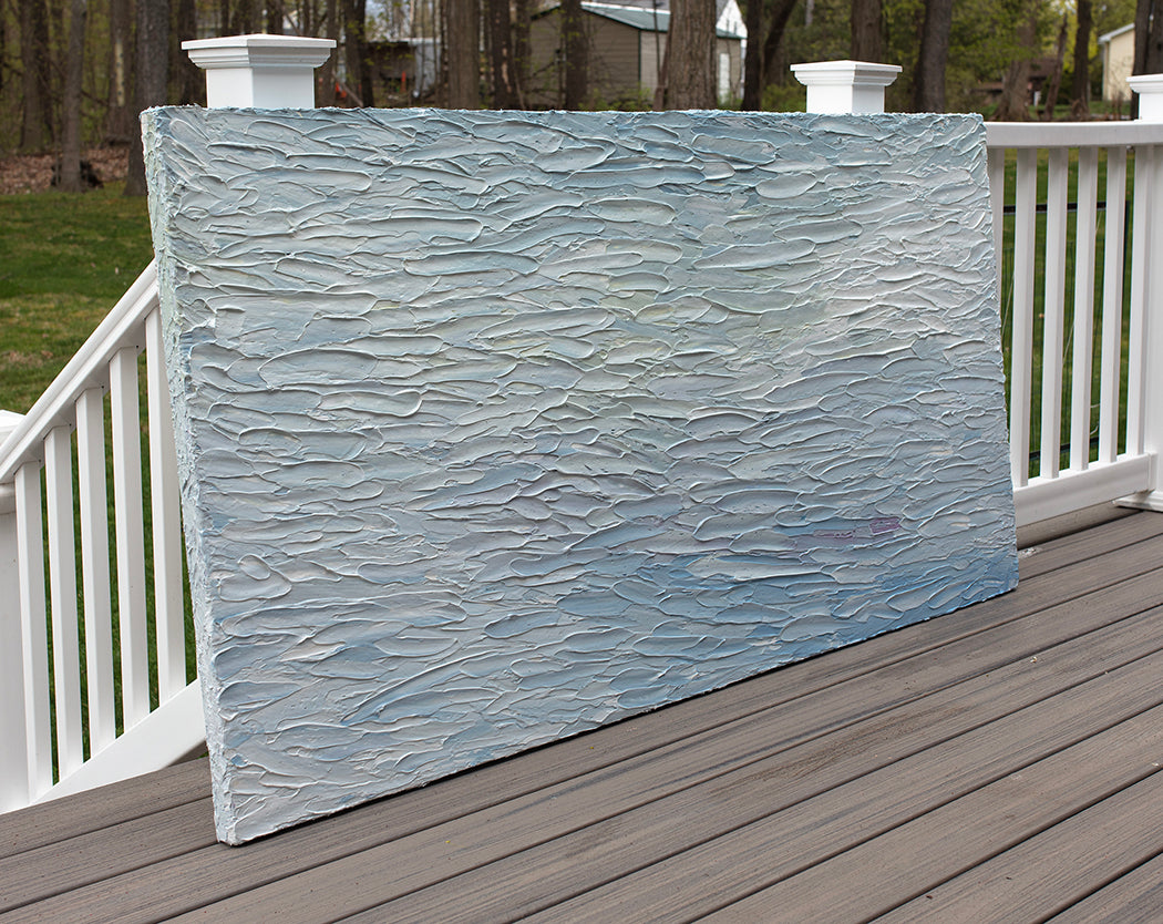 A abstract painting with blue textured brushstrokes is seen at an angle on a deck at the artists studio.