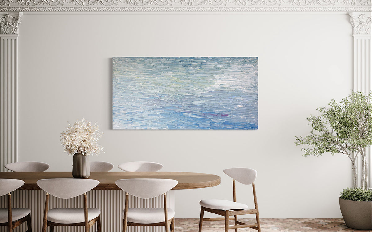A abstract painting with blue textured brushstrokes is seen hanging on a wall in a dining room over a table with white contemporary chairs and a potted tree to the right.