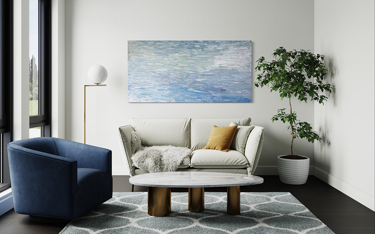 A abstract painting with blue textured brushstrokes is seen hanging on a wall in a family room over a beige loveseat with a potted tree to the right.