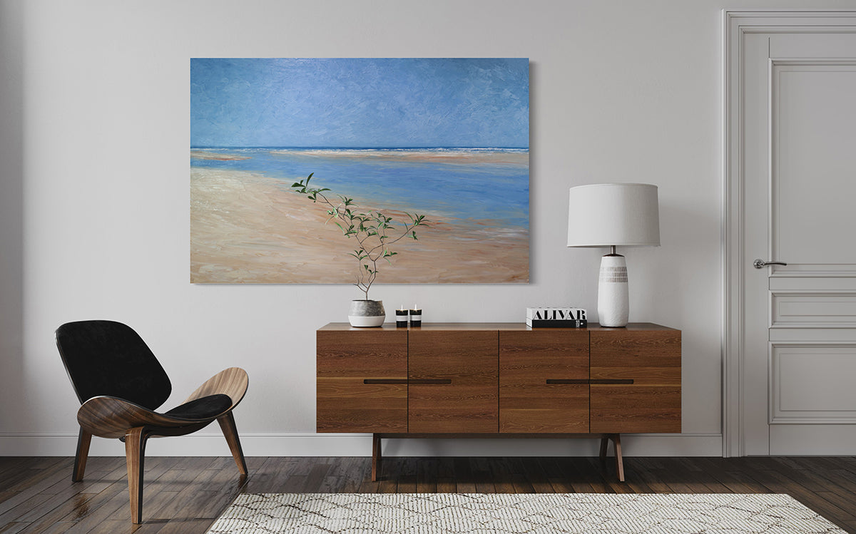 A textured blue and beige coastal painting of a beach hangs on a white wall above a mid-century credenza with a white lamp, books, candles and a plate on it. A chair sits to the left.