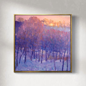 A painting of a snowy forest with a cool purple tone of the trees and the warm sun peaking behind the top framed in a gold floater frame hangs on a gallery wall.