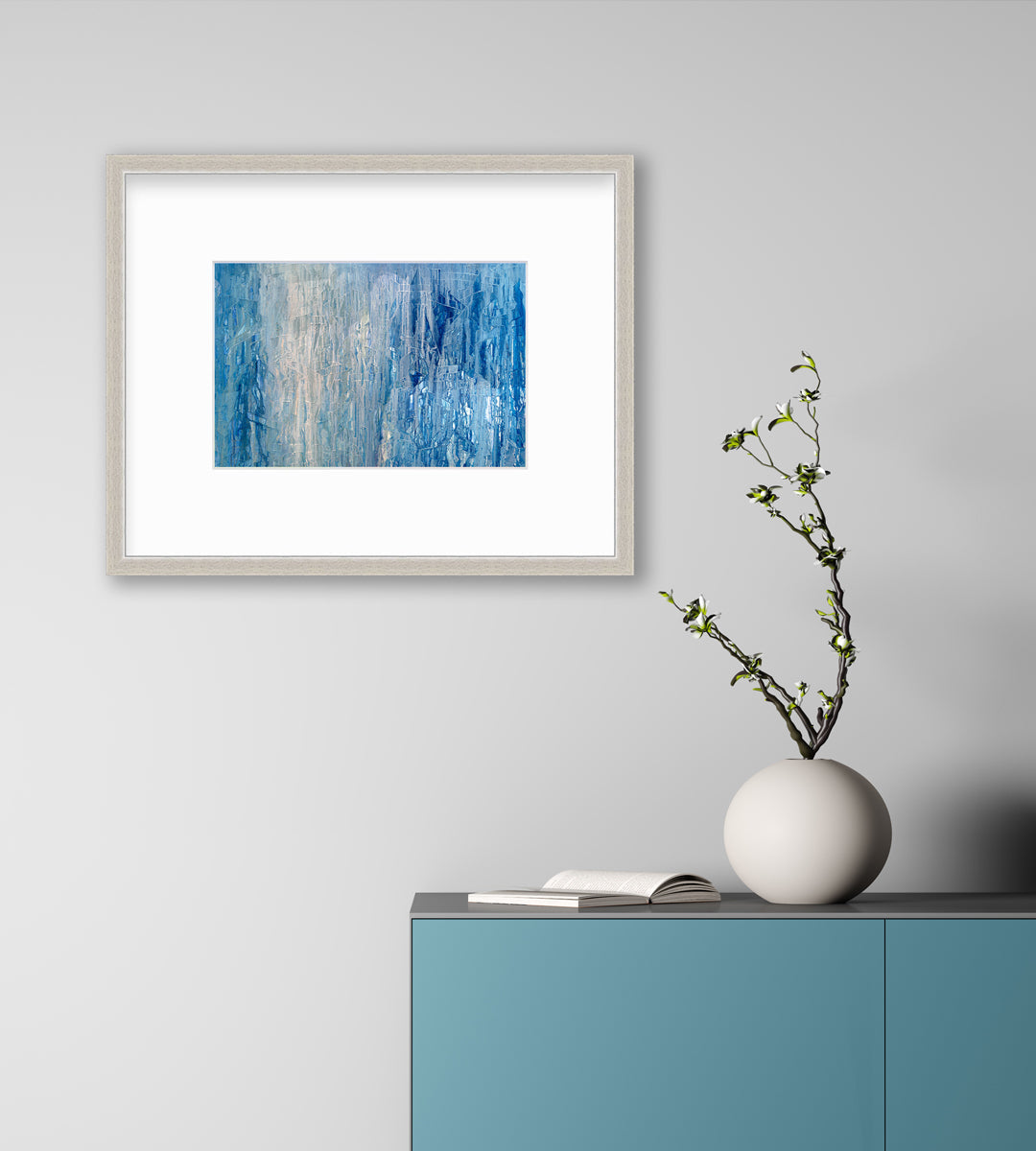 A blue, white and grey abstract print in a silver frame with a mat hangs on a white wall above a teal cabinet with a white round vase with greenery stems and an open book.