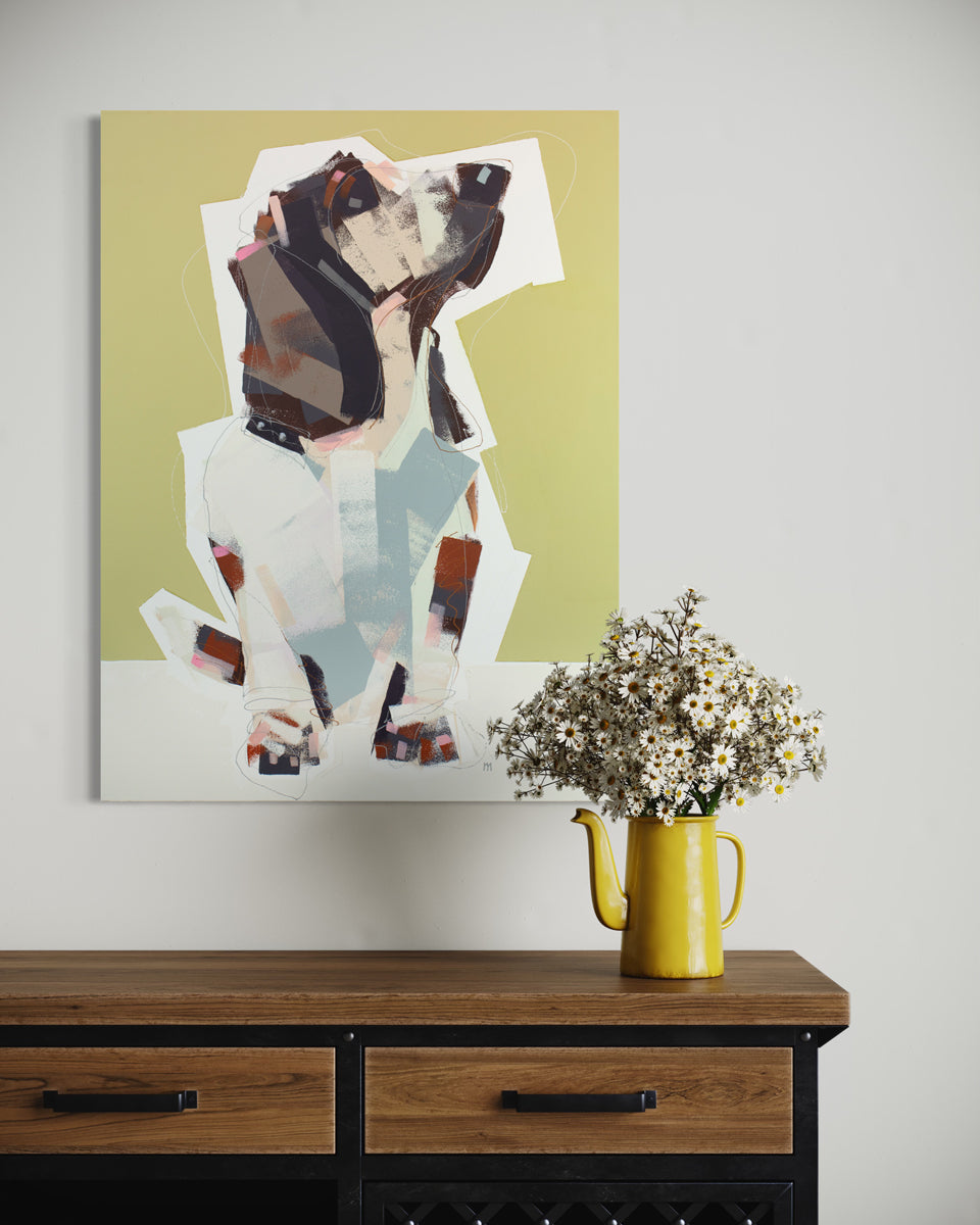 A yellow, white, and brown abstract painting of a dog hangs on a light wall above a wood console table with a flowers potted in a yellow watering can. 