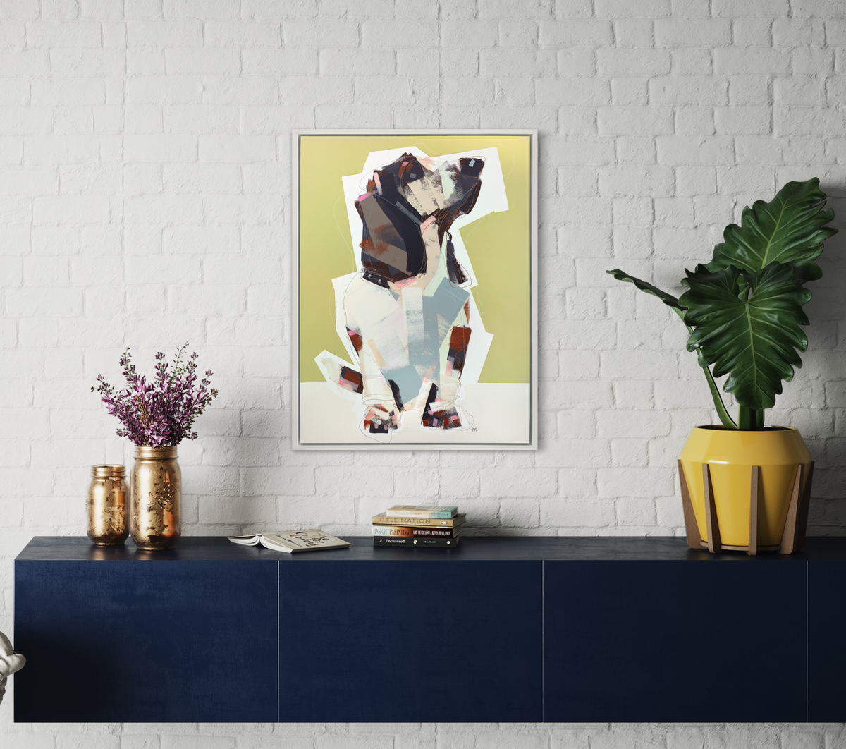 A chartreuse, navy, beige and light blue abstract dog print in a white floater frame hangs on a white brick wall above a floating blue console table with a stack of books, a medium sized potted plant and two gold mason jars, one with lavender flowers.