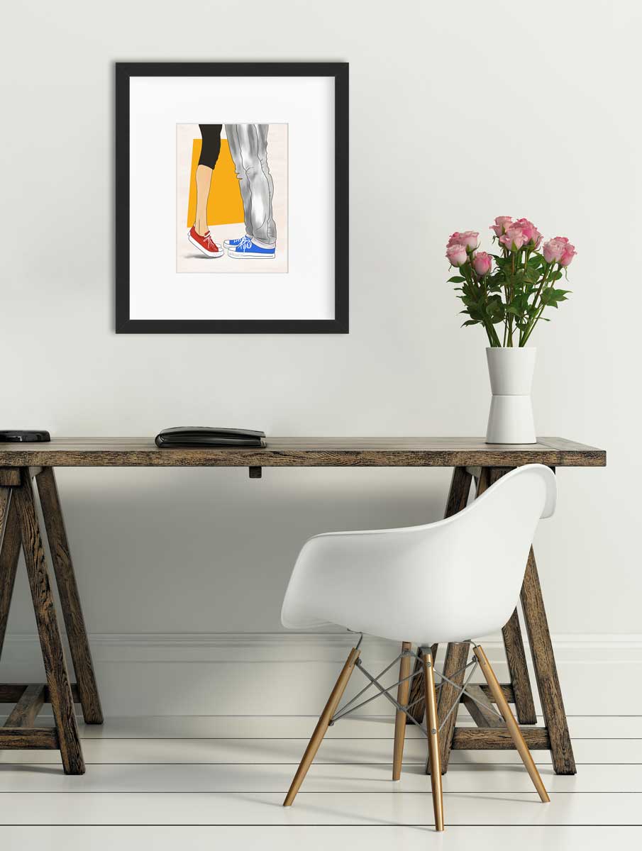 Art print with man and woman wearing Converse sneakers, matted and framed in a black frame and hanging on a white wall above a wood desk with a white plastic chair with wooden legs.
