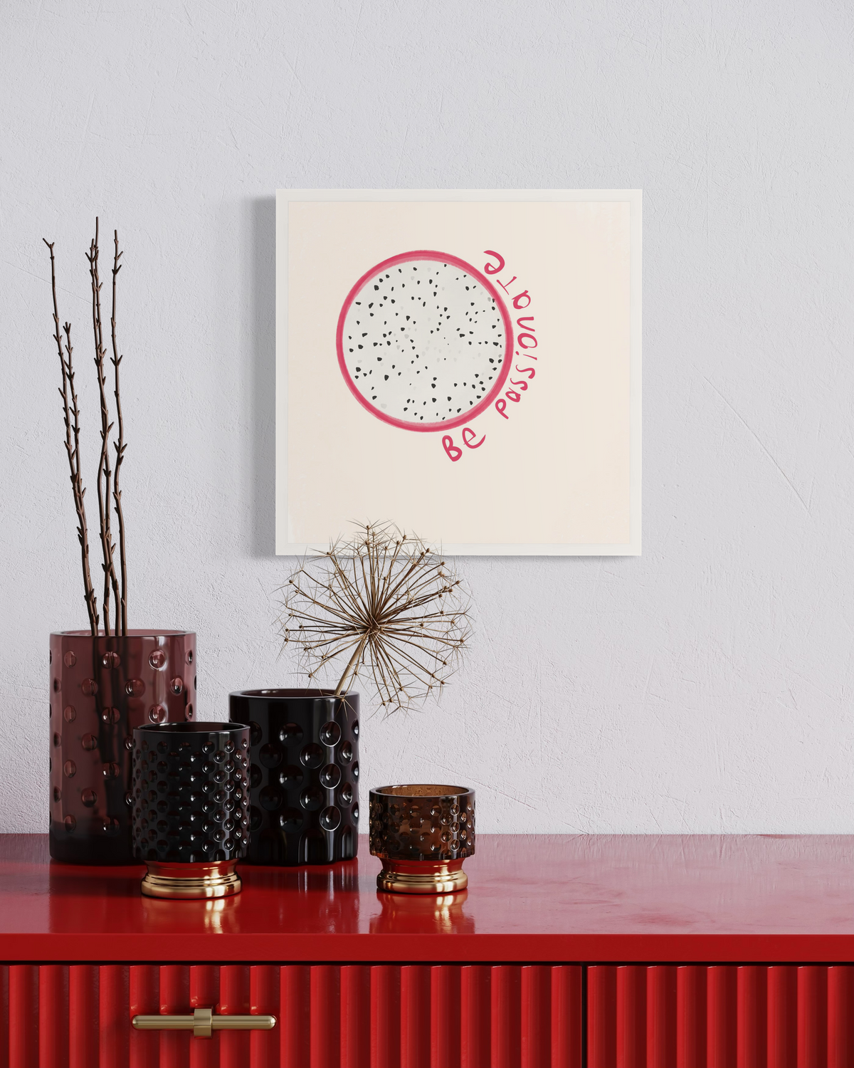 An art print of a minimalistic abstract fruit with the words "be passionate" in an unmatted white frame hangs on a white wall above a red desk with multiple red shaded vases with decorative stems.
