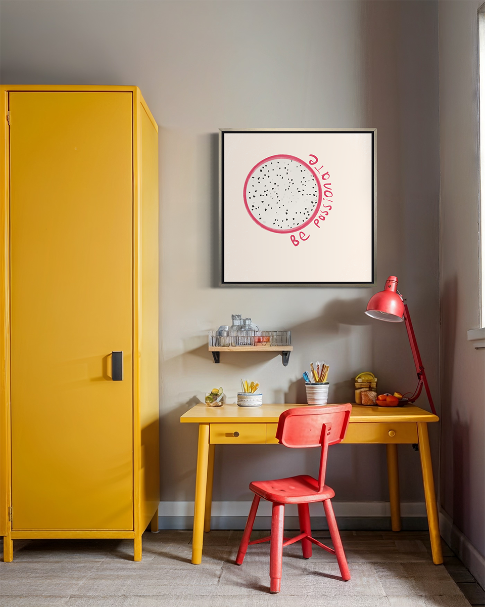 An abstract art print of a fruit framed in a silver floater frame hangs on a light wall above a yellow desk with cups of pencils, a red desk lamp and a red desk chair.