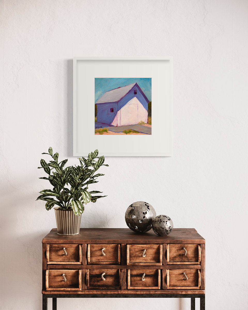 A blue, white and lavender contemporary barn print in a white frame with a mat hangs on a white wall above a distressed wood eight drawer cabinet with a potted plant and two grey sphere decorations.