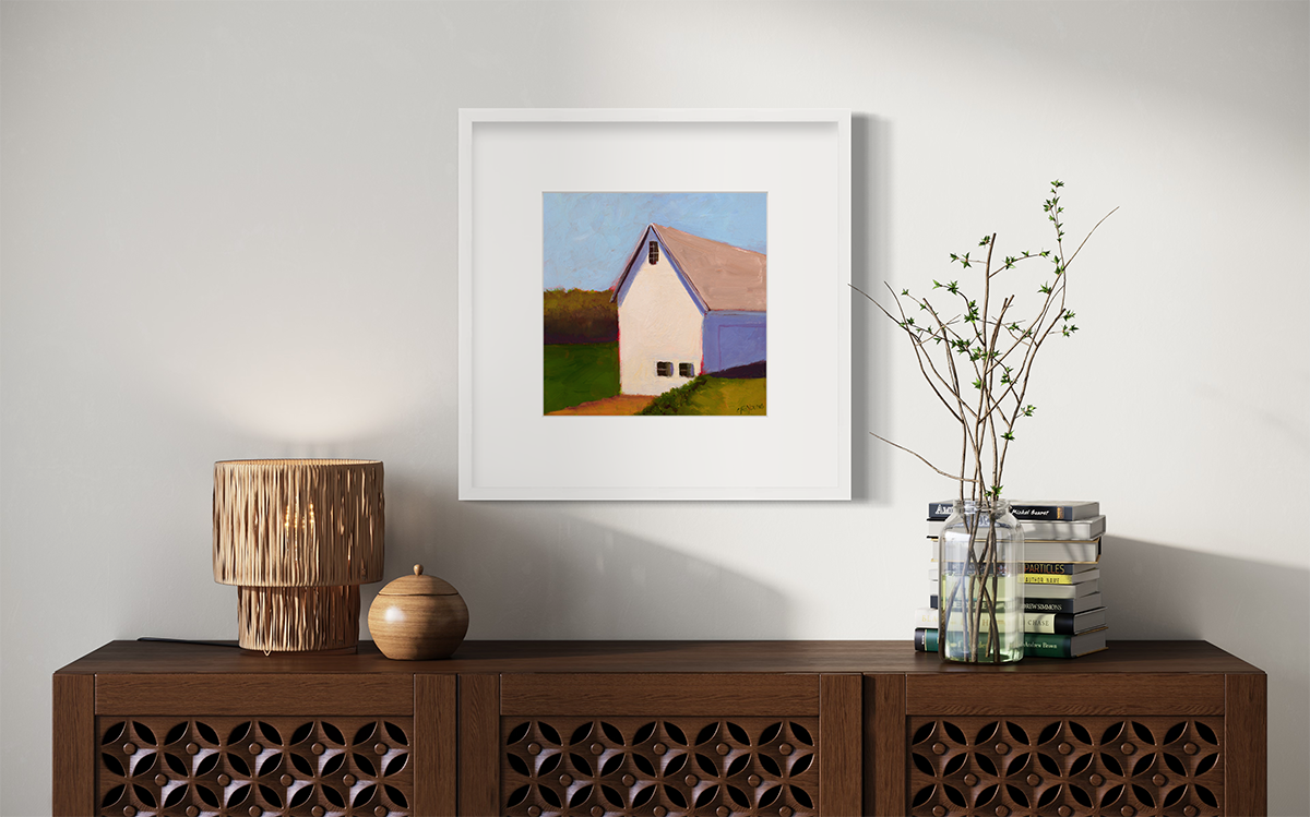 A blue, green, white and lavender contemporary barn print in a white frame with a mat hangs on a white wall above a dark wood credenza with a stack of books, a clear jar with stems and a small lamp.
