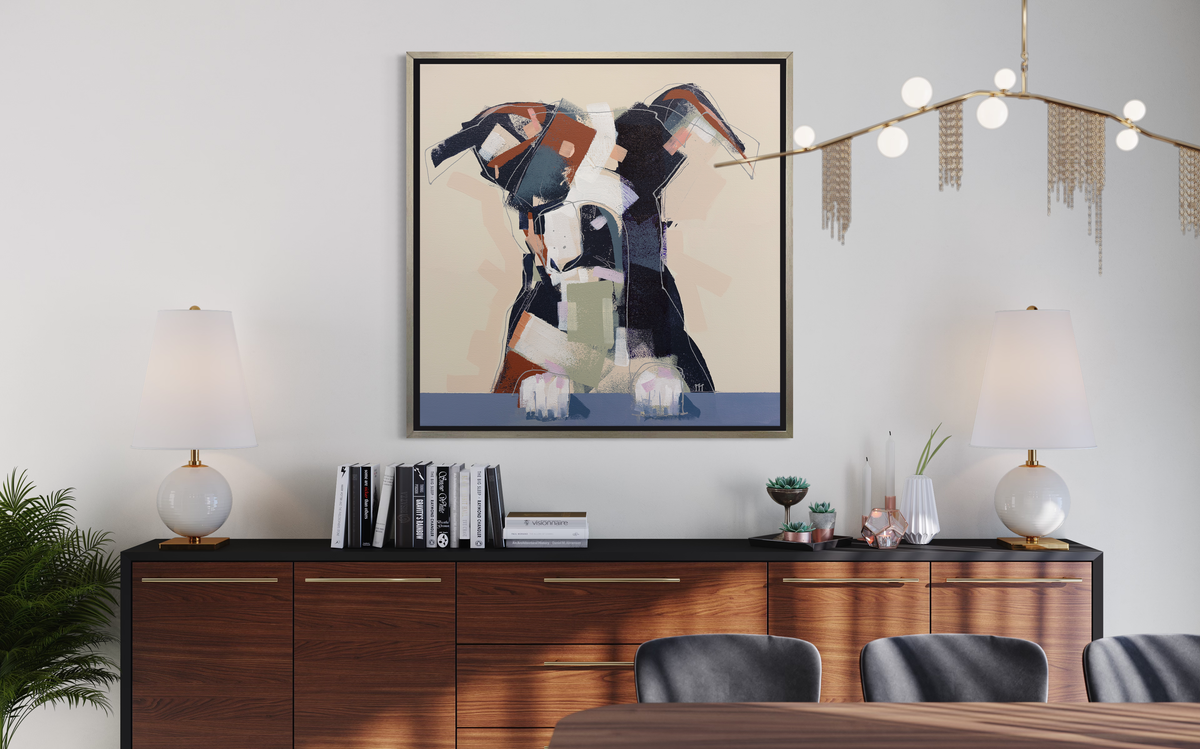A navy, brown, white and beige abstract dog print in a silver floater frame hangs on a white wall above a black and wood credenza with two matching white lamps, books and some decorative accessories.