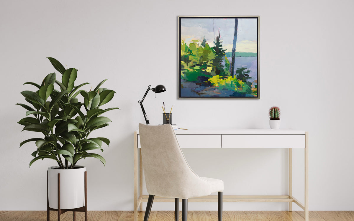 An abstract blue and green landscape print in a silver frame hangs on a white wall above a white desk and chair with a large potted floor plant to the left of it. 