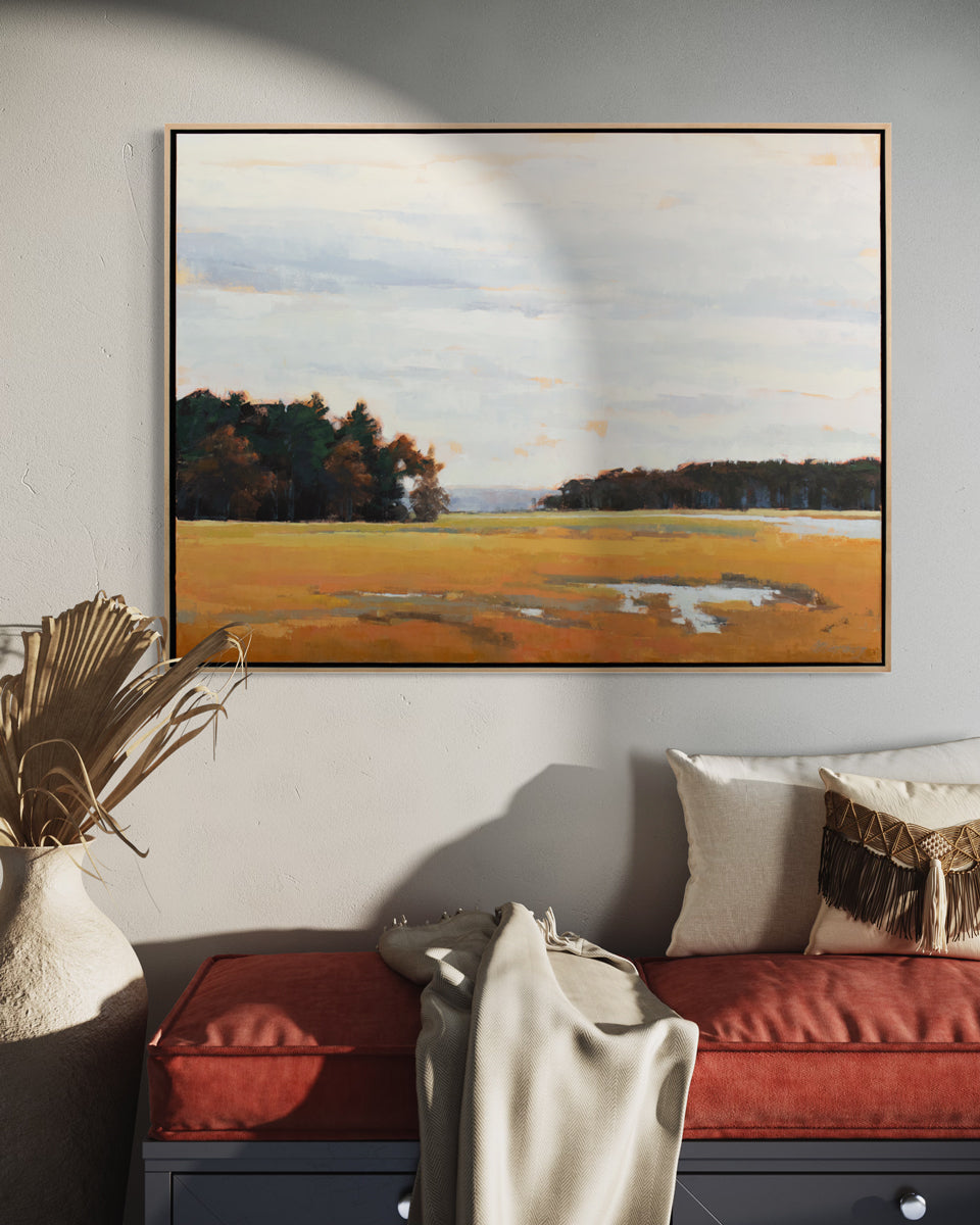 A light blue, dark green and shades of orange abstract landscape painting by Molly Doe Wensberg in a light wood floater frame hangs on a white wall above a grey bench with a rust red cushion, neutral pillows and a white throw blanket. 