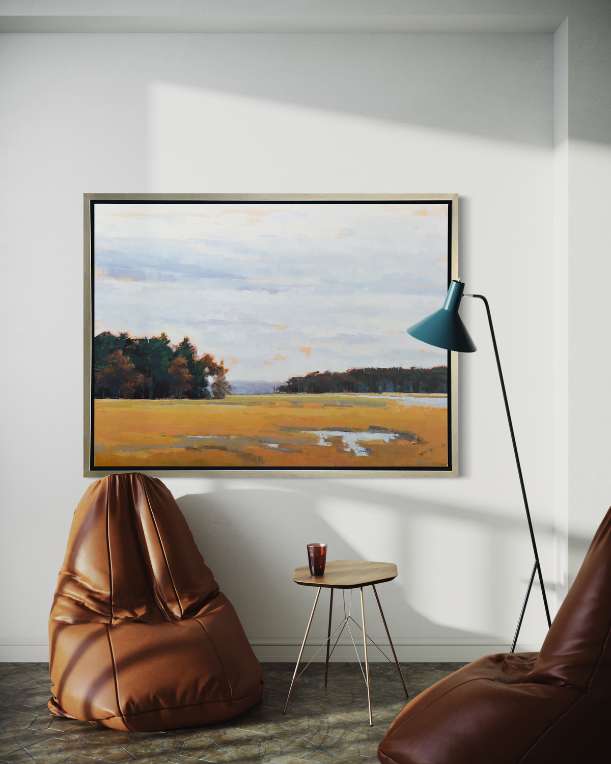 A landscape print in a silver floater frame hangs on a white wall above an oblong brown leather bean bag chair, a small side table and a teal floor lamp.