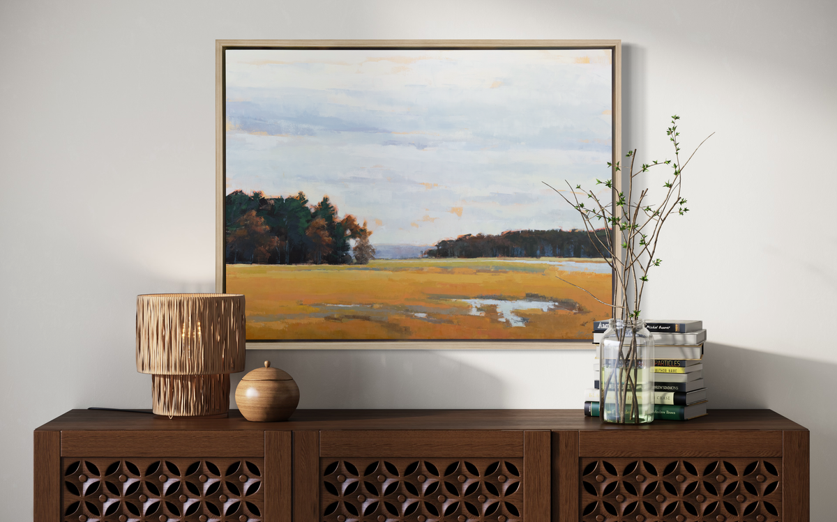 A landscape print in a white oak wood floater frame hangs on a white wall above a dark wood credenza with decorative accessories.