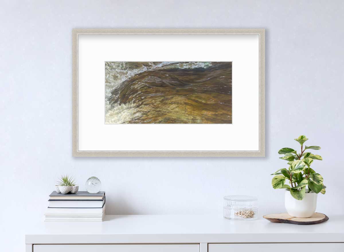 A brown, beige and white abstract river print in a silver frame with a mat hangs on a white wall above a white countertop with a stack of books, a clear round container with gold accessories and a small potted plant.