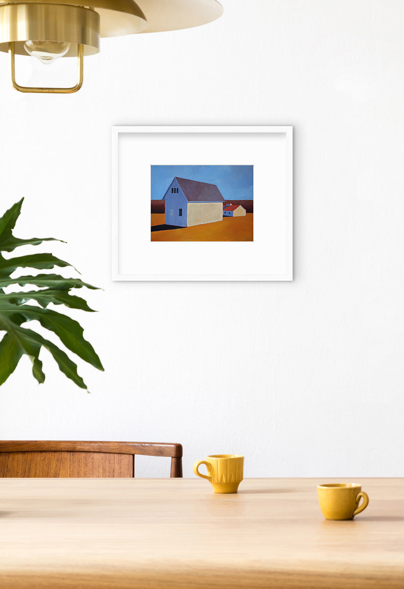 A blue and orange contemporary barn print in a white frame with a mat hangs on a white wall above a wood table with two yellow tea cups sitting on top and a modern gold chandelier hanging above.