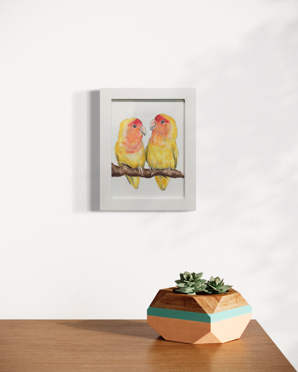 An illustration of yellow and orange birds sitting on a branch together framed in a white frame hangs on a white wall above a wood table surface with a geometric planter holding succulents. 