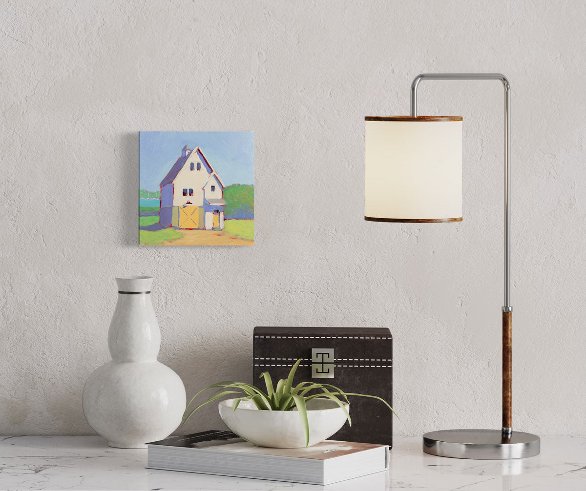 A small green, blue, violet, and yellow painting of a house hangs on a textured wall above black and white table decor and a small modern lamp. 