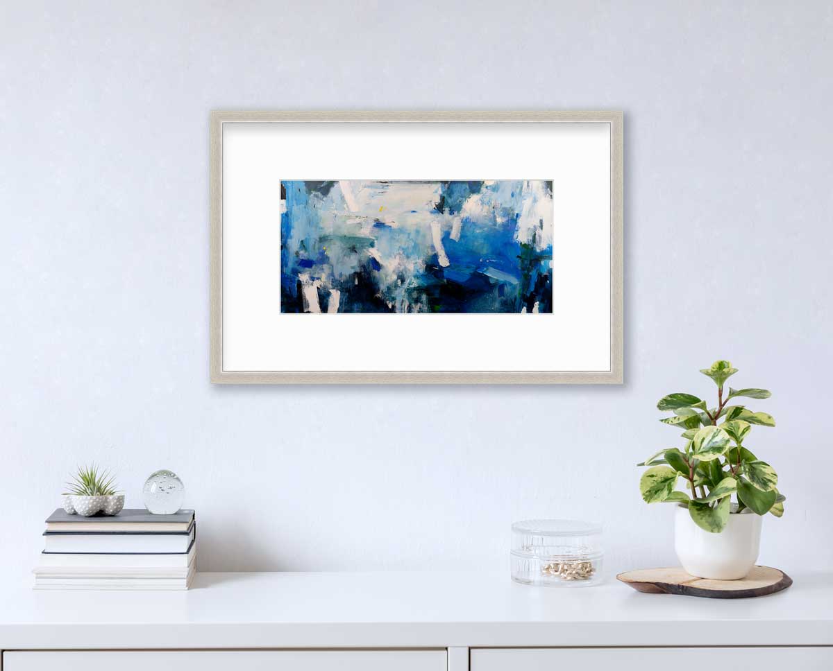 A blue and white abstract print in a silver frame with a mat hangs on a white wall above a white surface with a stack of books, a clear round container with gold accessories and a potted plant.