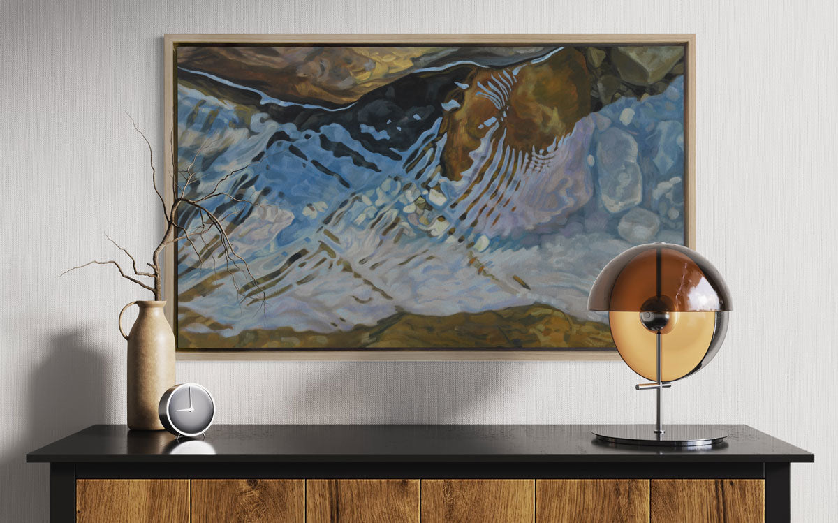 A print of water reflecting over river rocks framed in a wood frame hangs on a white wall above a modern black and wood console table with modern table decor. 