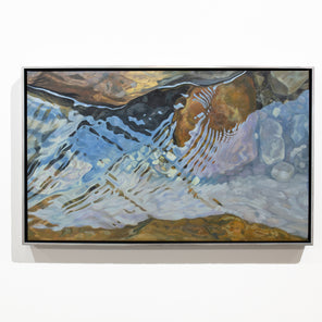 A naturalistic painting of river rocks framed in a silver frame hangs on a white wall. 