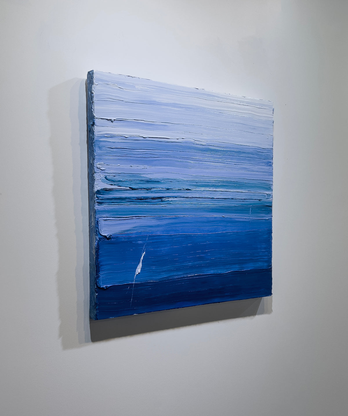 A blue and white textured abstract painting by Teodora Guererra hangs on a white wall, side view.
