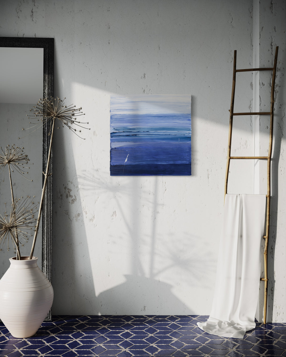 A blue and white textured abstract painting by Teodora Guererra hangs on a white wall.  To the right is a natural wood decorative ladder with a white fabric draped over it. to the left is a wall mirror with a dark ridged frame leaning on the wall and a large white ceramic pot with decorative sticks in front. The floor below is a dark blue tile in a geometric pattern with grey grout.