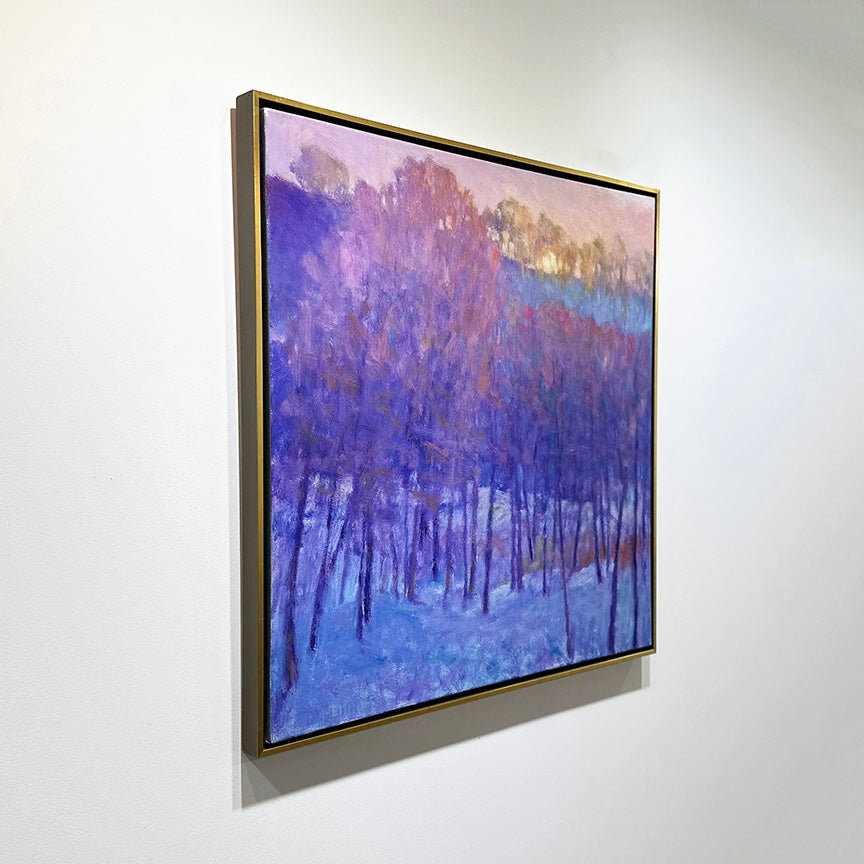 A painting of a snowy forest with a cool purple tone of the trees and the warm sun peaking behind the top framed in a gold floater frame hangs from a side view on a gallery wall.