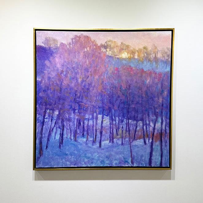 A painting of a snowy forest with a cool purple tone of the trees and the warm sun peaking behind the top framed in a gold floater frame hangs on a gallery wall.