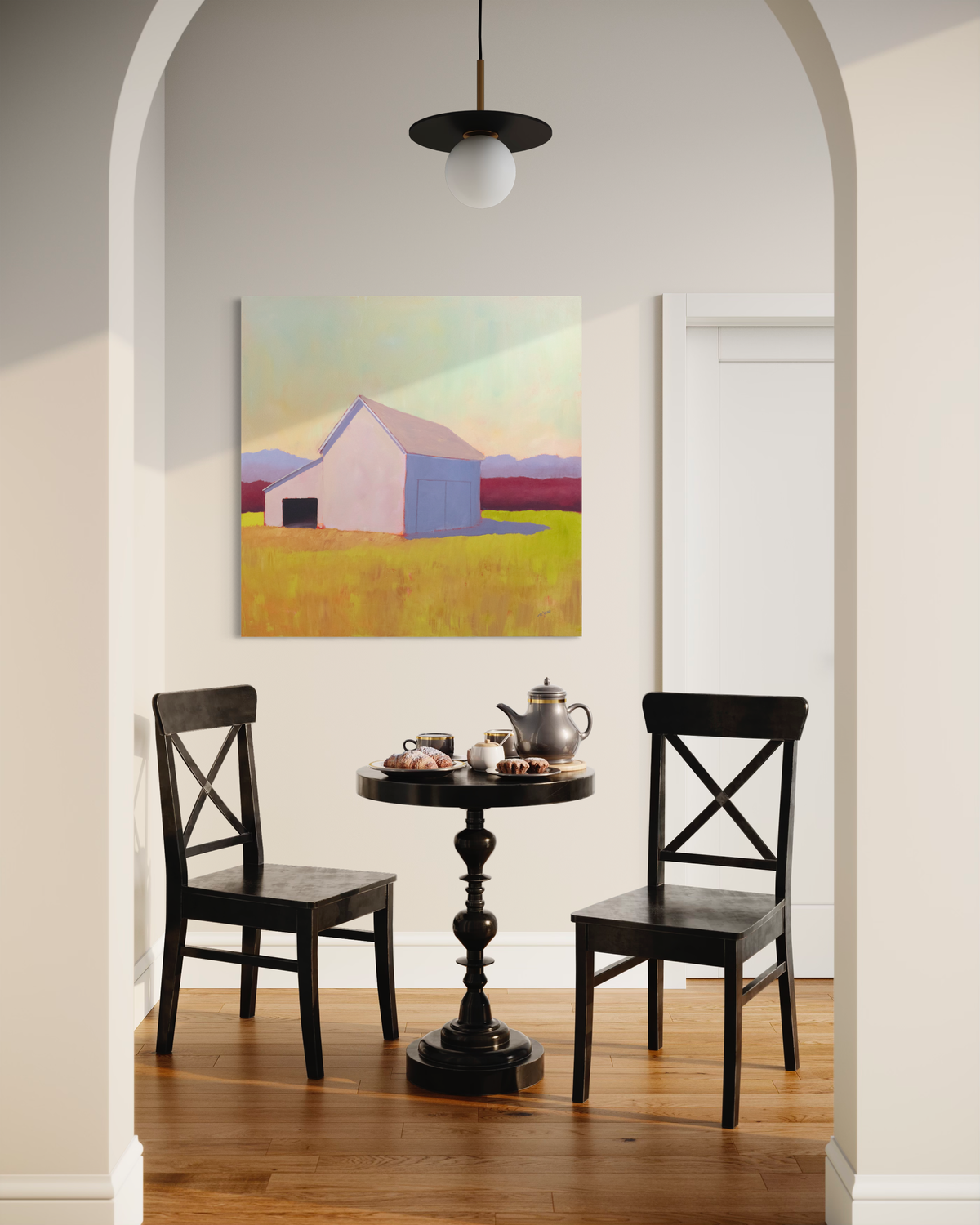 A landscape painting of a blush lavender barn with a tinted yellow sky, lavender and raspberry colored hedges and a chartreuse and orange foreground by Carol Young hangs on a cream wall above two dark wooden chairs and a matching pedestal table with a tea set and a baked goods. 