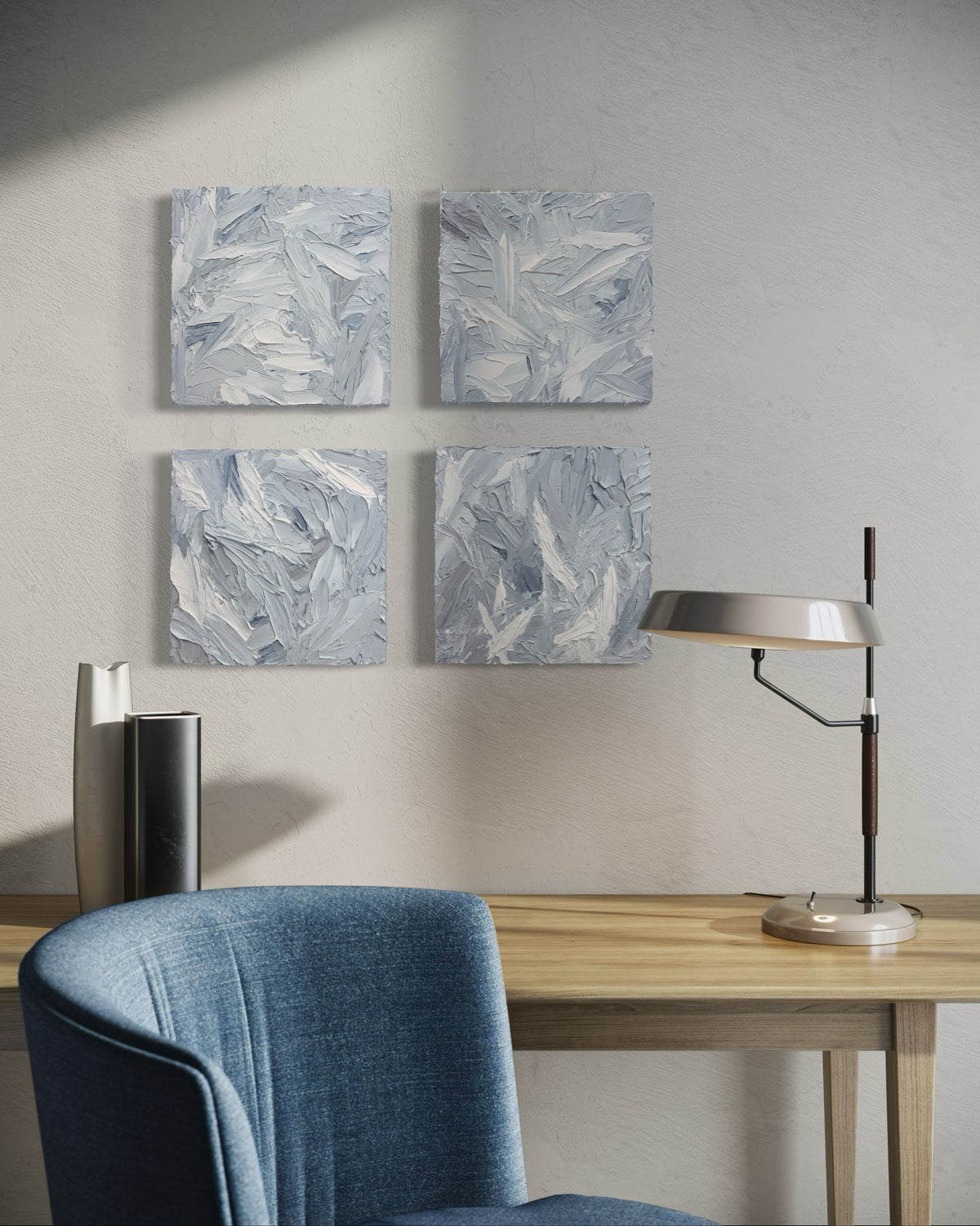 Four 12x12 inch paintings thickly painted with palette knife strokes in blue, white and grey hang on a wall over a maple desk with a blue chair.