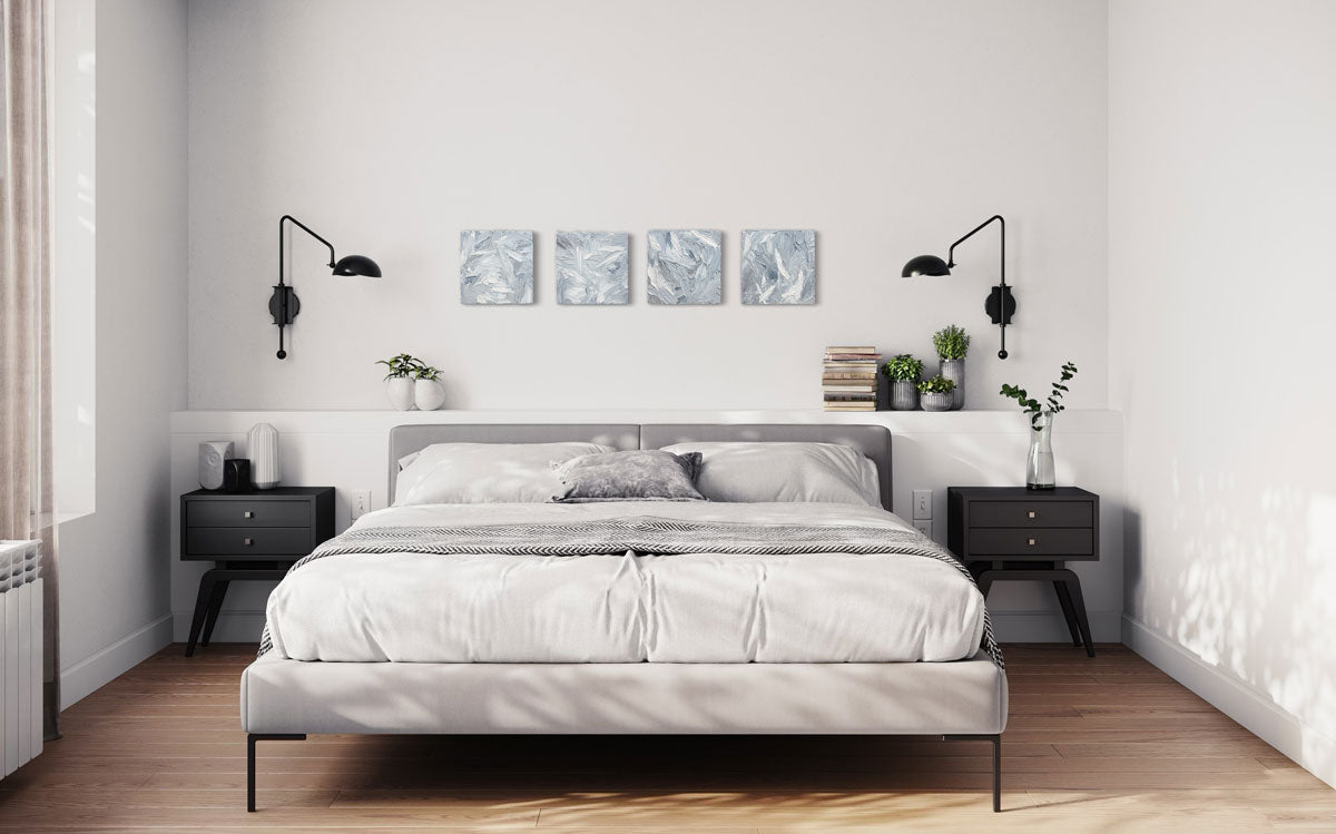 Four 12x12 inch paintings thickly painted with palette knife strokes in blue, white and grey hang on a wall over a bed in horizontal display.
