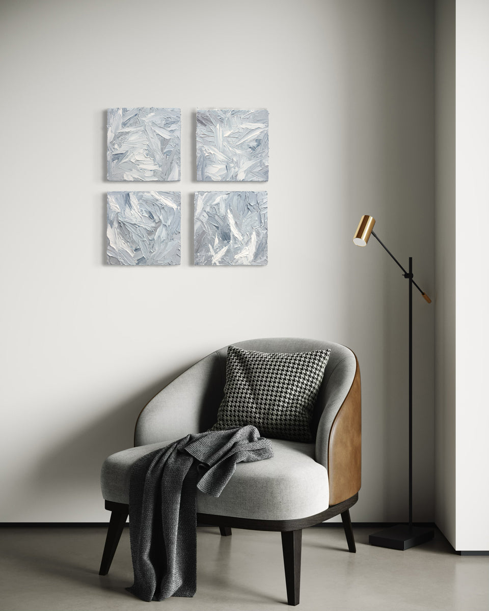 Four 12x12 inch paintings thickly painted with palette knife strokes in blue, white and grey hang on a wall over a grey chair.