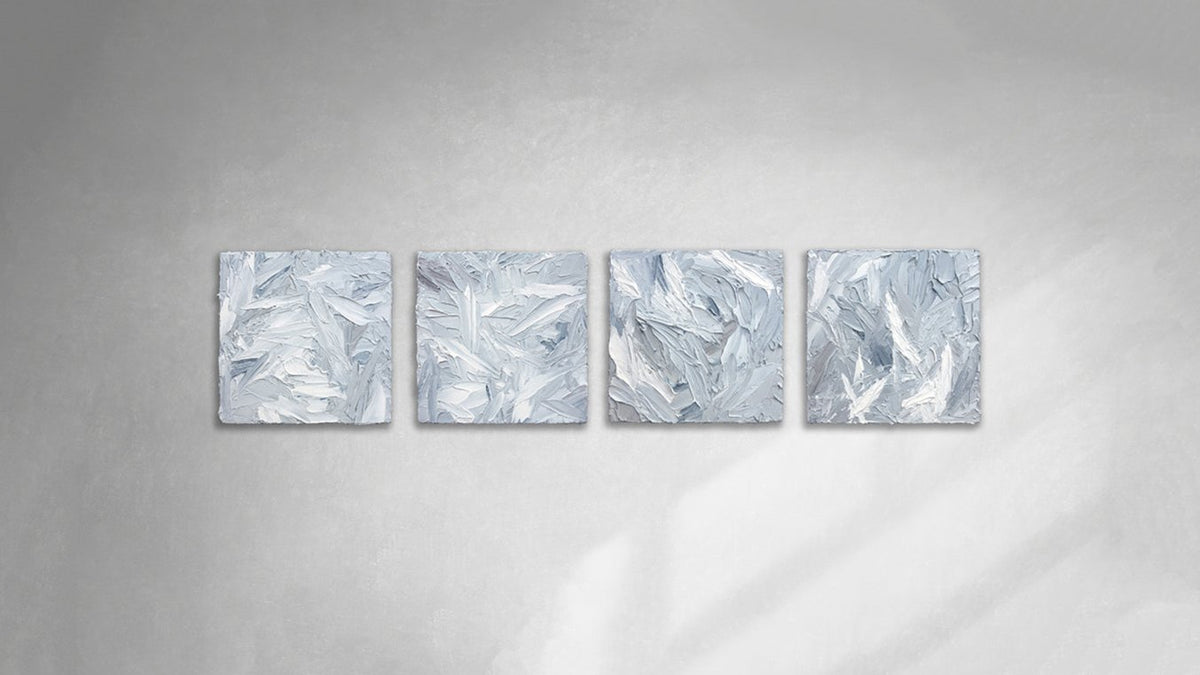 Four 12x12 inch paintings thickly painted with palette knife strokes in blue, white and grey hang on a wall in a horizontal display.