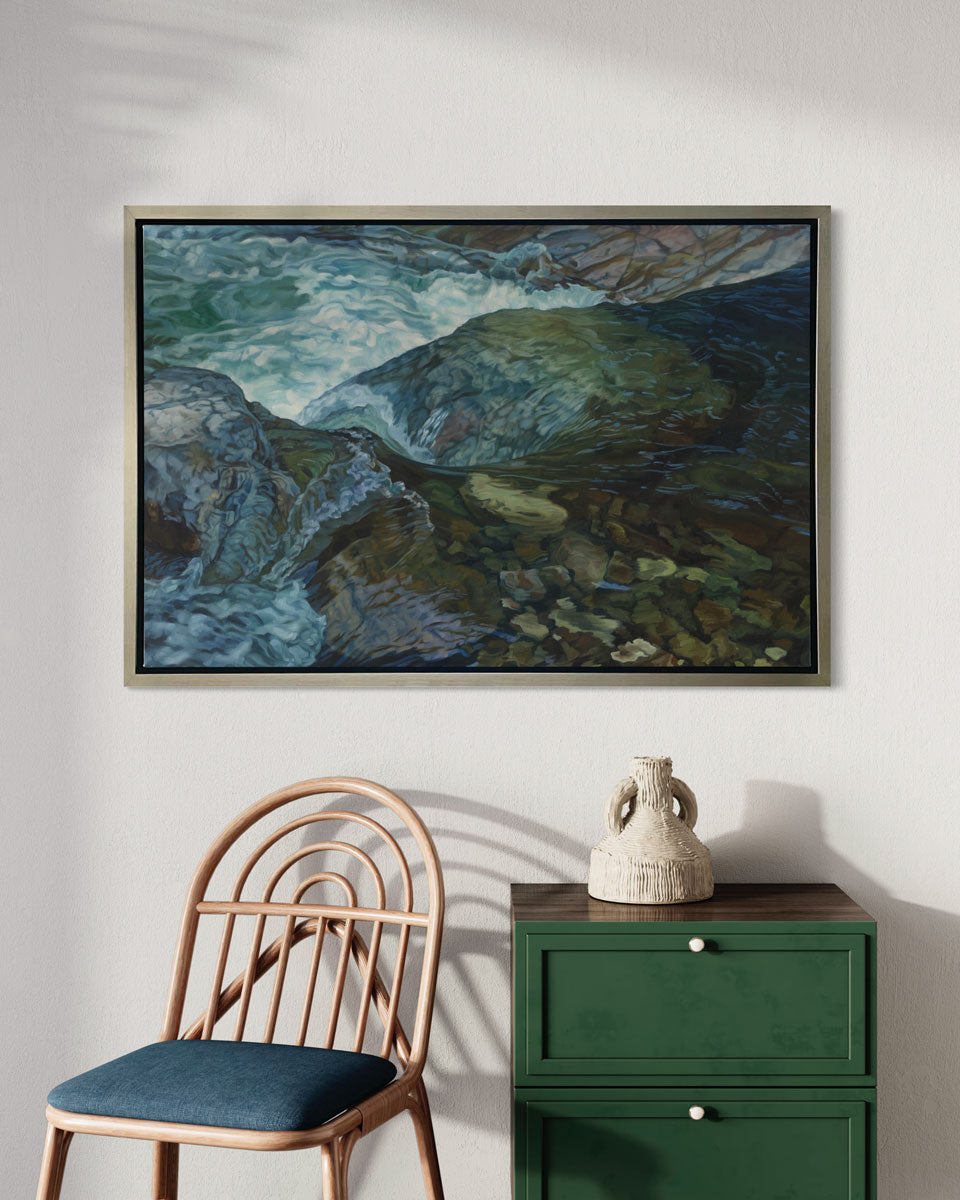 An art print of a riverbed scene framed in a silver frame hangs on a white wall above a green end table and a wood armchair with a blue seat cushion. 