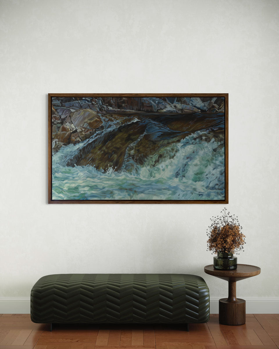 A print of river water streaming over rocks framed in a wood frame hangs on a white wall above a modern green leather bench and a wooden end table with a vase full of flowers. 