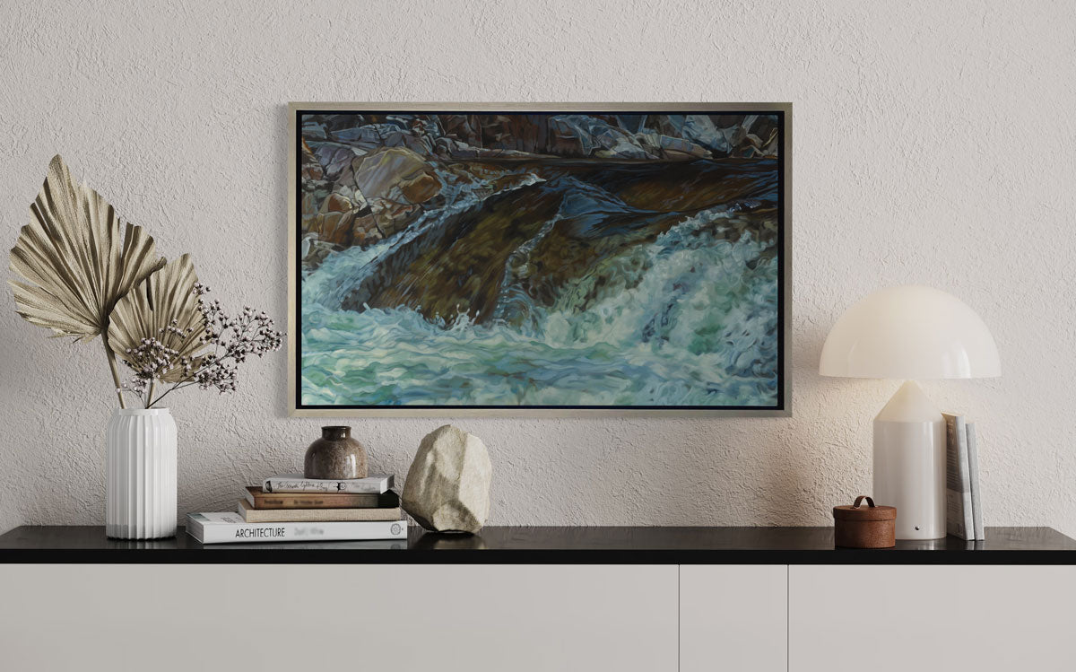 An art print of water running over river rocks framed in a silver frame hangs on a white wall above a black and white console table with modern table decor. 