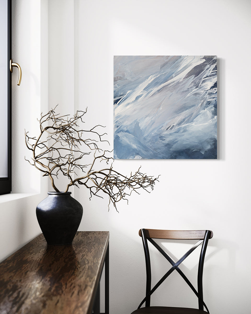 A white, grey and blue abstract painting by Teodora Guererra hangs on a white wall above a chair with a ceramic pot with dried branches on a shelf under a window to the left.