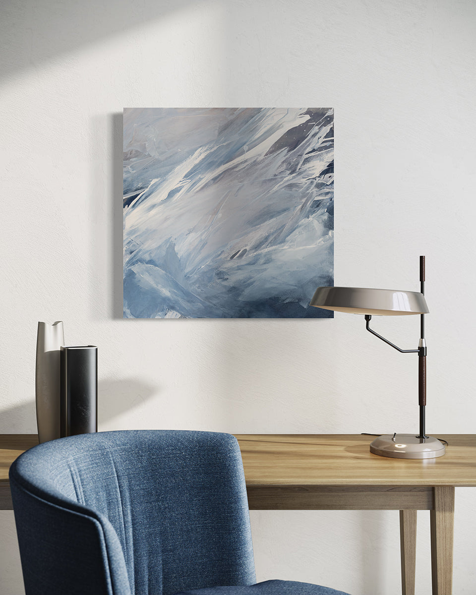 A white, grey and blue abstract painting by Teodora Guererra hangs on a white wall above a desk with a lamp and 2 ceramic vases.  In front of the desk is a blue chair. 