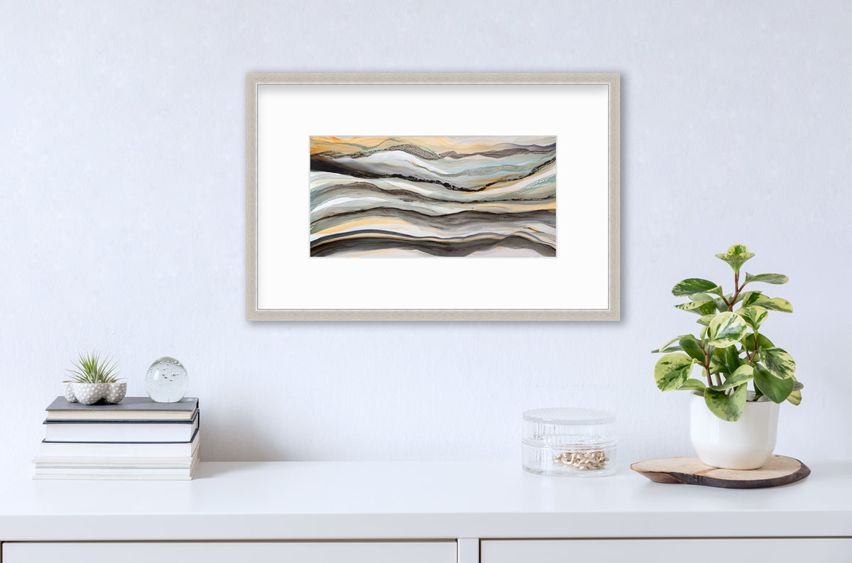 A black, white, orange and teal abstract print in a silver frame with a mat hangs on a white wall above a white surface with a stack of books, a round clear container with gold accessories and a potted plant.