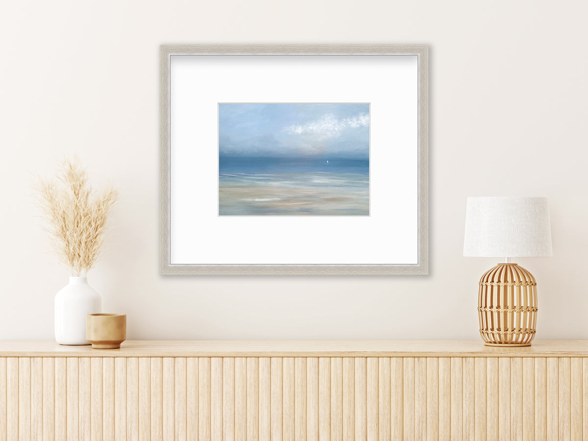 A light blue, white and beige abstract seascape print in a silver frame with a mat hangs on a white wall above a wood surface with a table lamp, a beige decorative cup and a white vase with wheat stems.