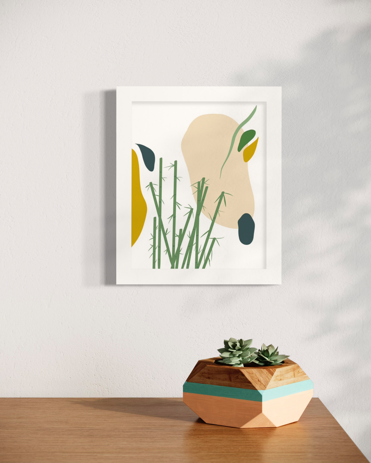 An abstract yellow, beige, blue, and green botanical art print in an unmatted white frame hangs on a white wall above a wood surface with a painted wood planter with a succulent arrangement.