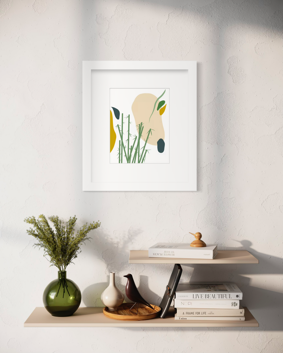 An abstract yellow, beige, blue, and green botanical art print in a white frame with a mat hangs on a white wall above two natural wood floating shelves with stacks of books, a green vase with greenery and decorative accessories.