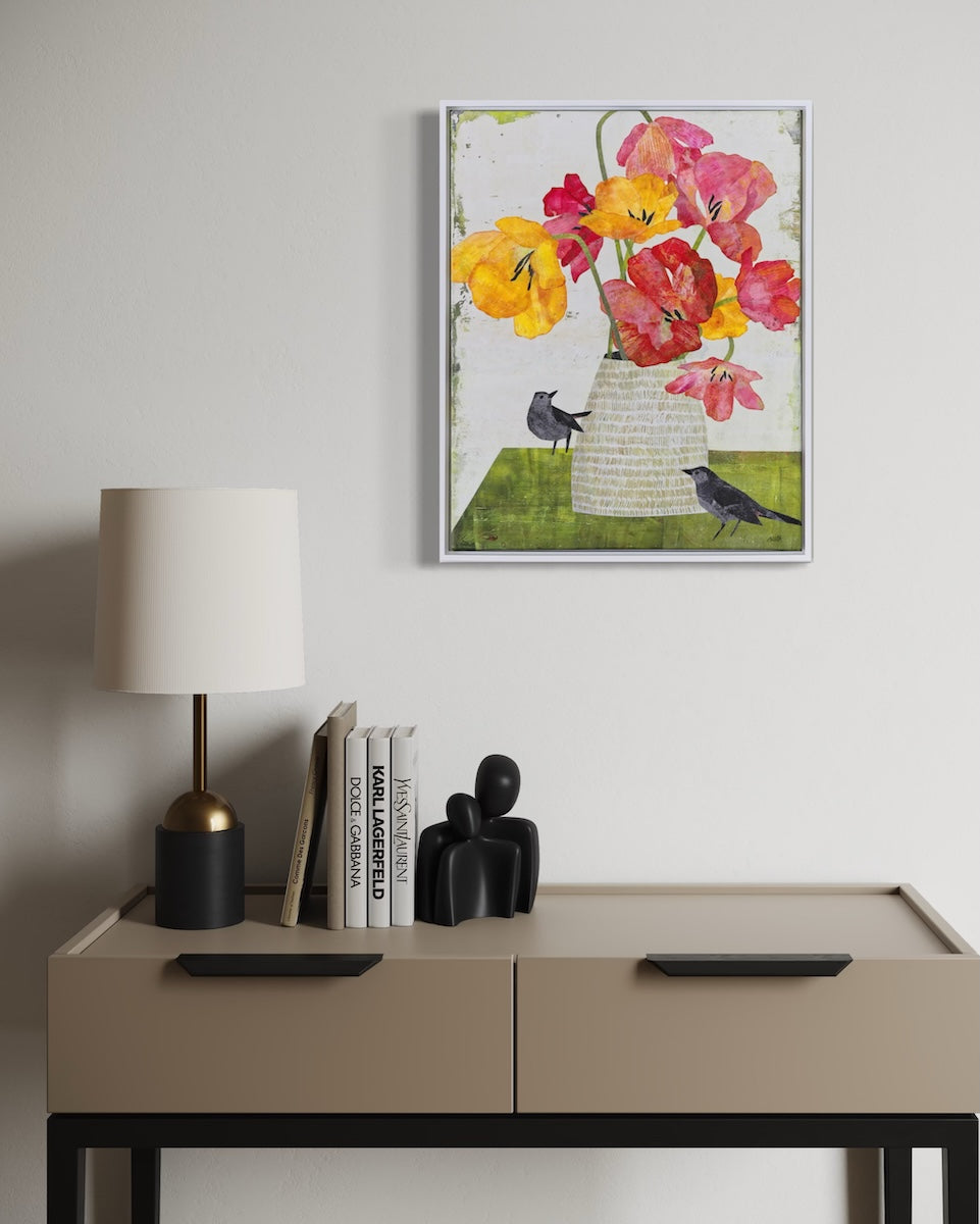 A framed collage featuring a floral bouquet with yellow, pink, and red flowers in a textured vase, displayed on a light gray wall above a beige console table with a lamp and books.