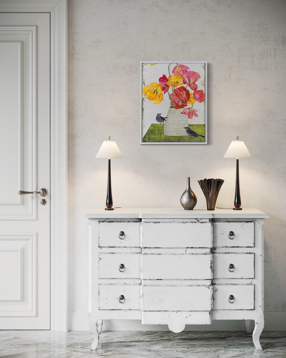 A framed floral collage featuring pink, yellow, and orange flowers with small birds, displayed on a textured light gray wall above a distressed white wooden dresser in a styled room setting.
