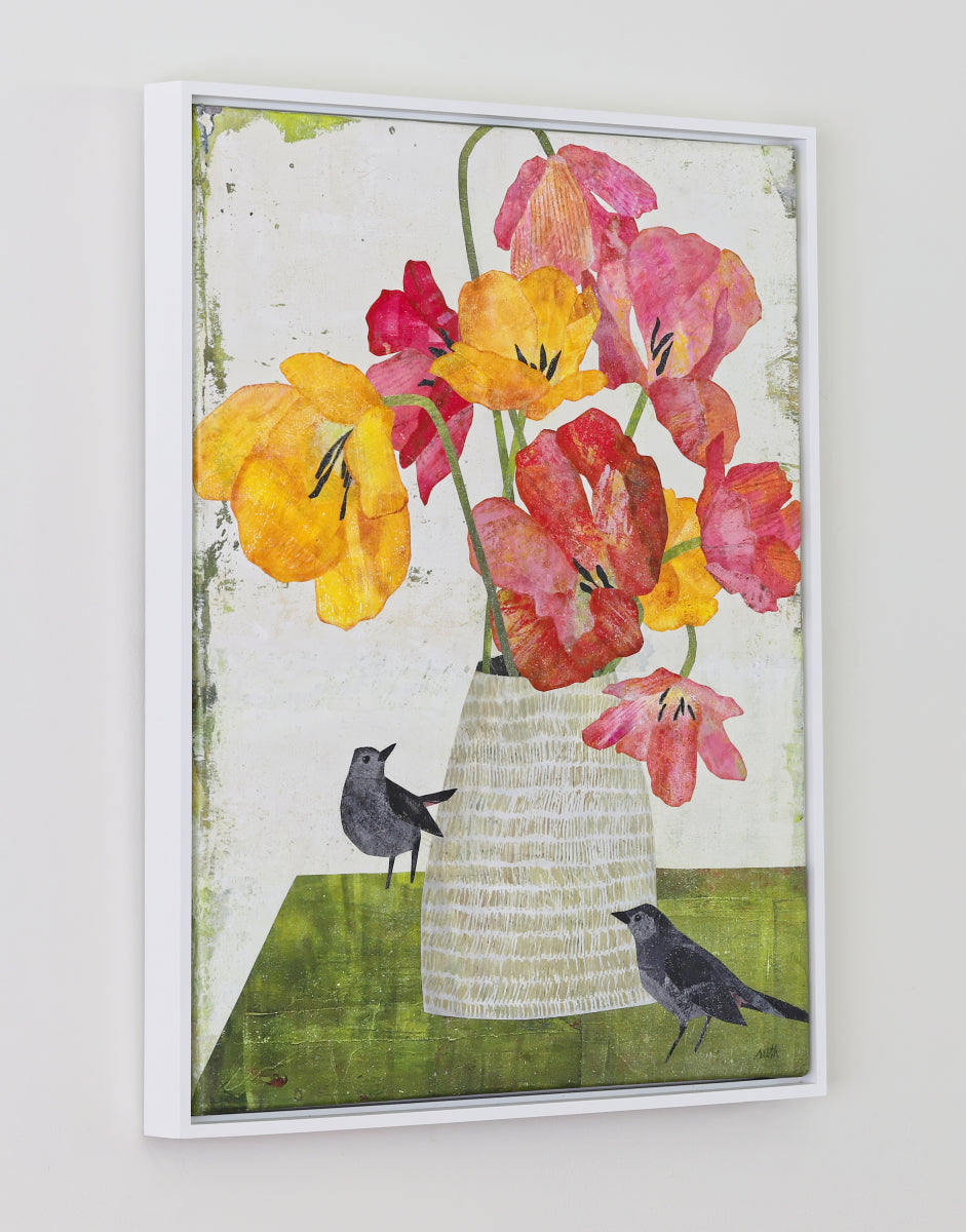 A framed collage featuring a bouquet of yellow, red, and pink flowers in a white vase, with two small black birds on a green surface against an off-white background. The artwork is framed in a clean white frame and displayed on a neutral light gray wall.