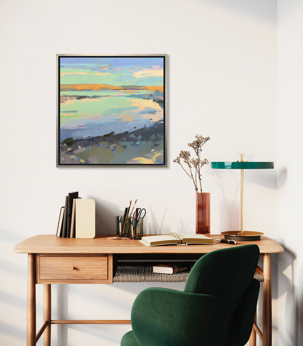 A colorful abstract landscape print framed in a silver frame hangs on a white wall above a wooden desk with green lamp and armchair. 