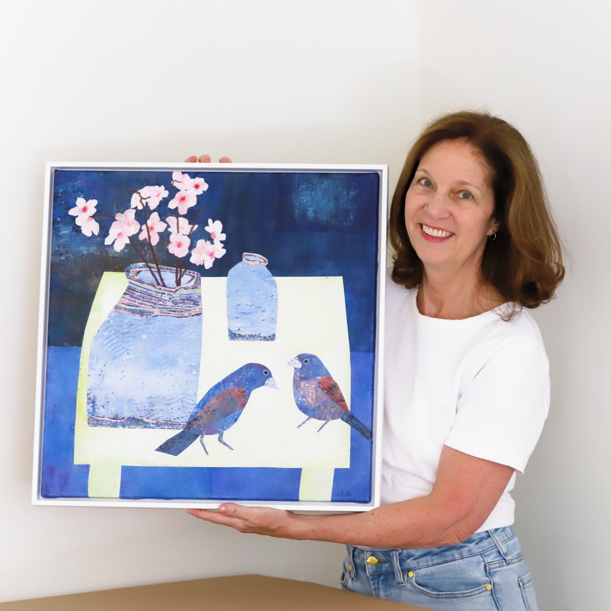 A smiling woman in a white t-shirt and jeans holds up a framed artwork depicting blue birds, blue vases, and pink blossoms against a dark blue background.