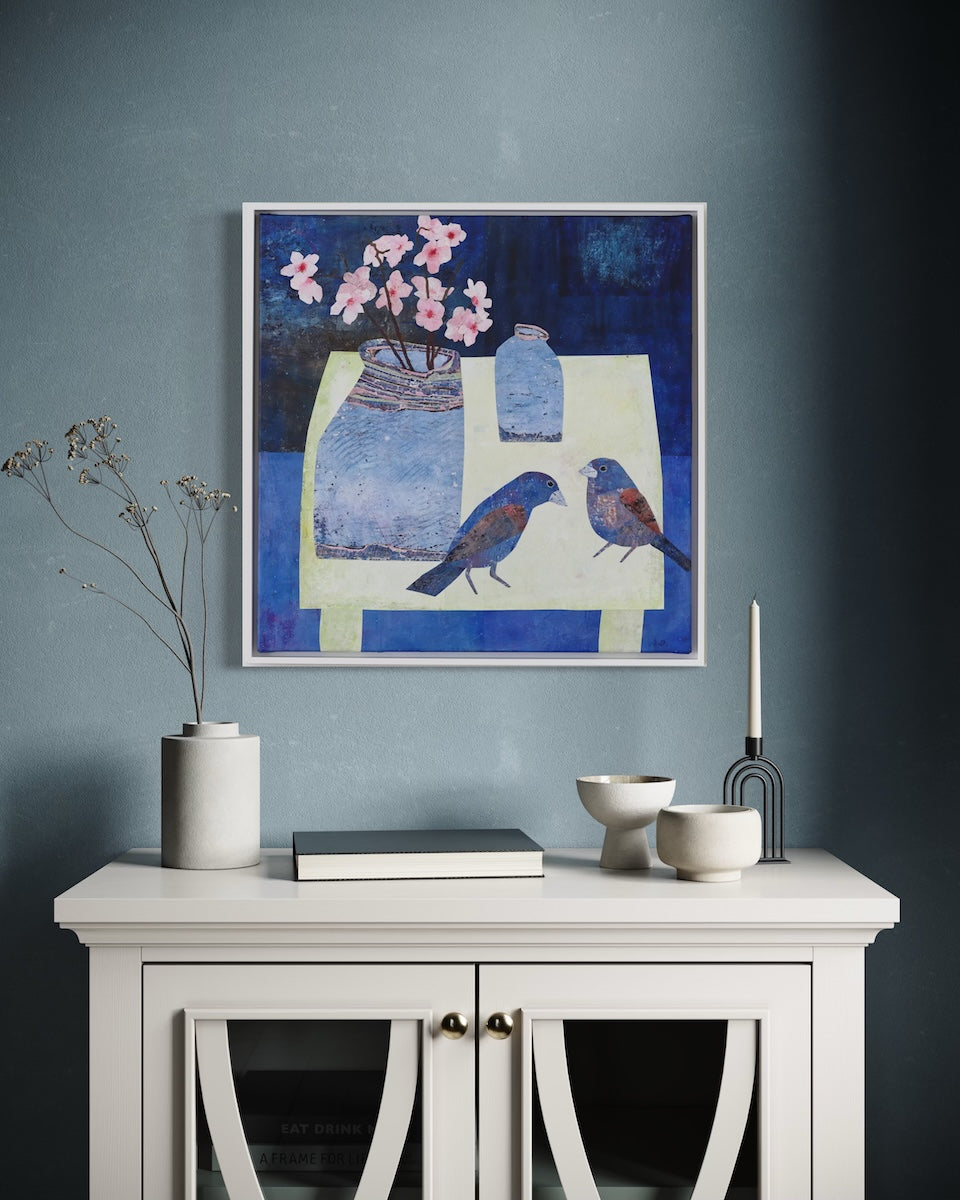 A framed square collage depicting two blue birds and pink flowers on a table, styled on a dark blue wall above a white cabinet with decorative ceramic vases and bowls.
