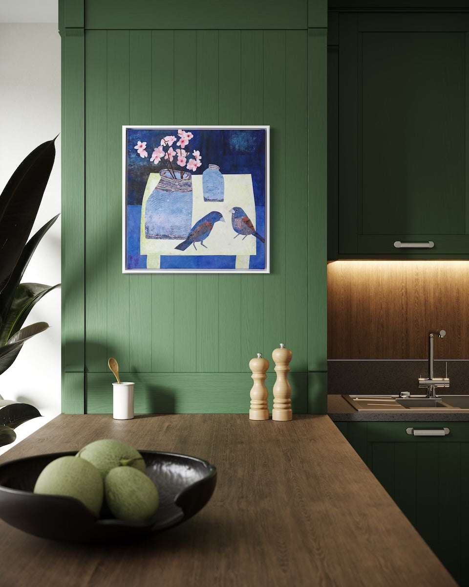 A framed square collage painting depicting blue birds and pink flowers hangs on a dark green paneled wall above a wooden kitchen counter with a bowl of fruit.